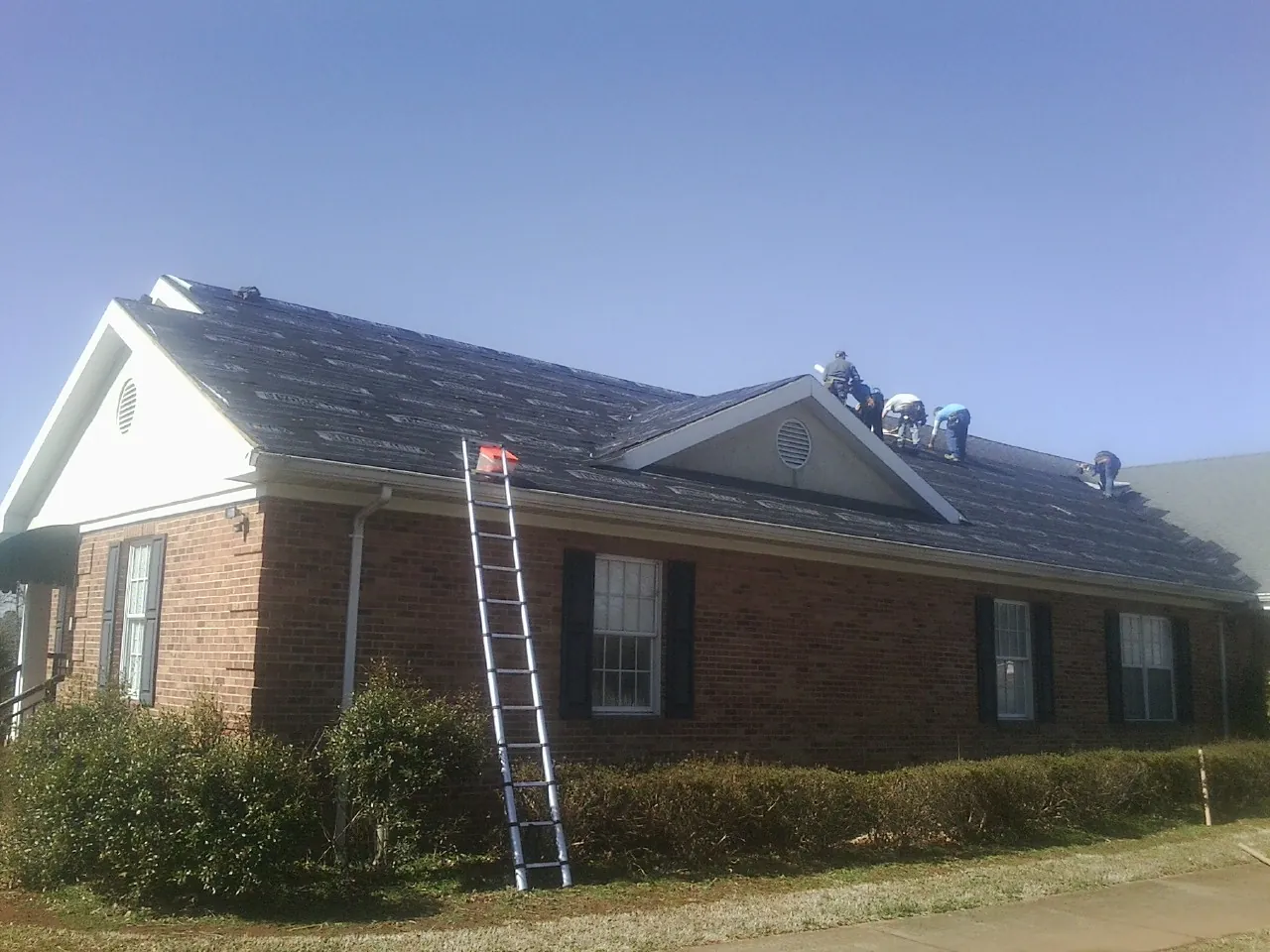 Crew tearing shingles off a brick ranch roof with a ladder set — Emergency Roof Repair work by our crew in Mazomanie