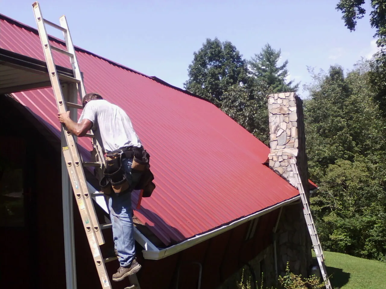 Roofer on a ladder beside a red metal roof and stone chimney — Soffit & Fascia Repair by Roofing Brothers in Mazomanie